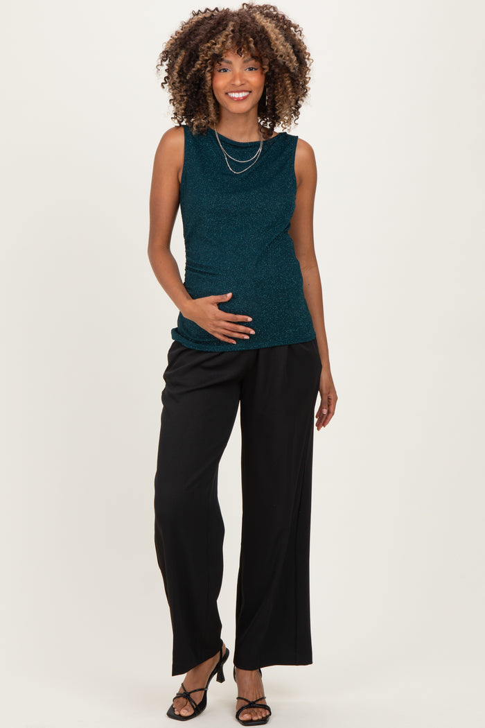 woman wearing a teal sparkly maternity top and black wide-leg pants, gently holding her baby bump