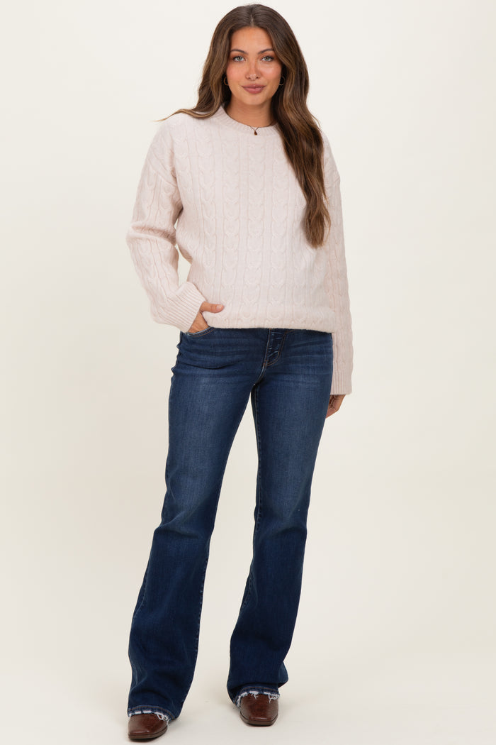 woman wearing a light pink cable-knit maternity sweater and dark blue flare jeans, posing with one hand in her pocket