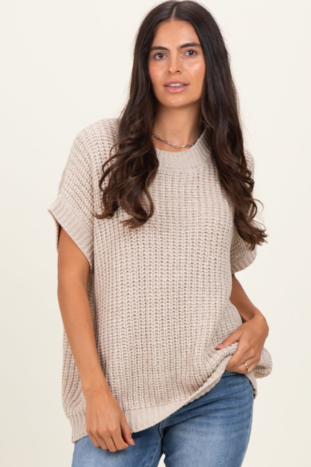 woman wearing a beige short sleeve knit sweater and blue jeans, standing with one hand in her pocket