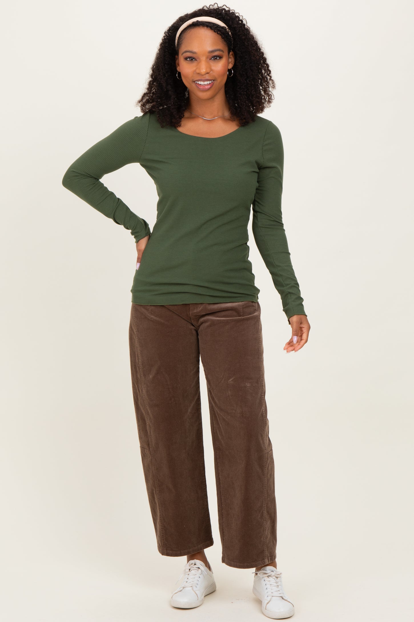 woman wearing a dark green long sleeve fitted top and brown wide leg corduroy pants, standing with one hand on her hip