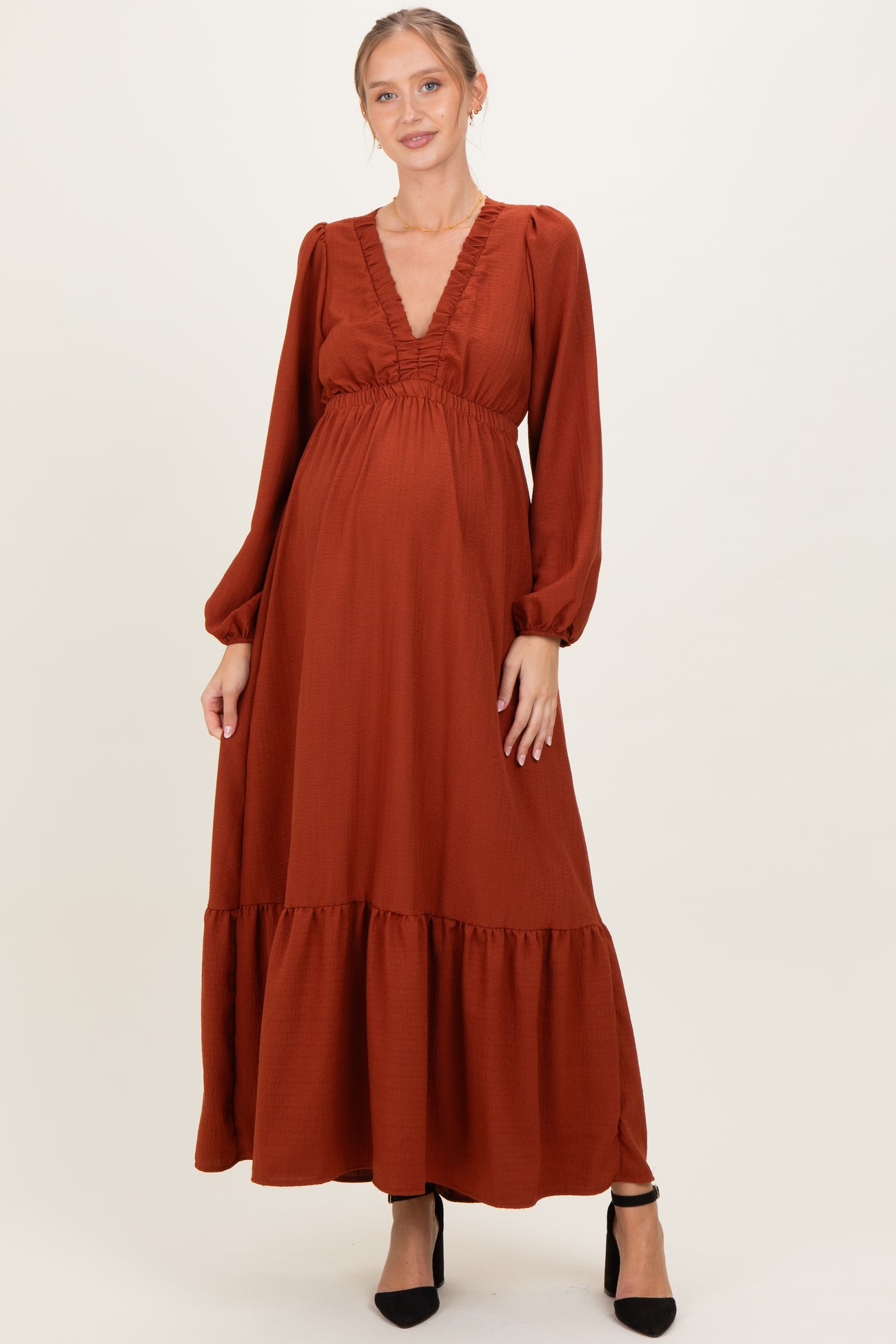 woman wearing a rust-colored maternity maxi dress with a ruffled neckline and long sleeves, highlighting her baby bump