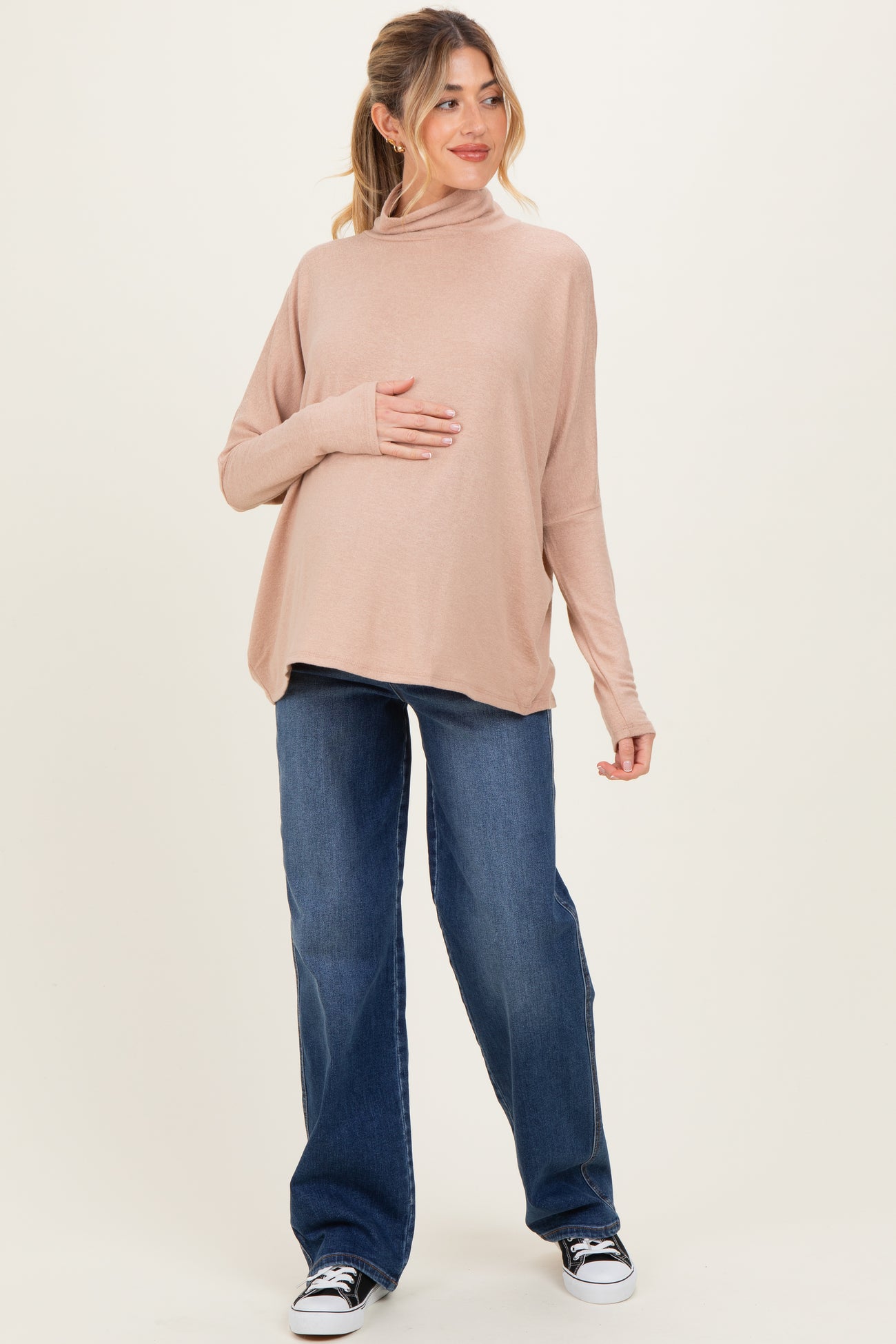 woman wearing a light pink long sleeve maternity top and blue wide-leg jeans, holding her baby bump