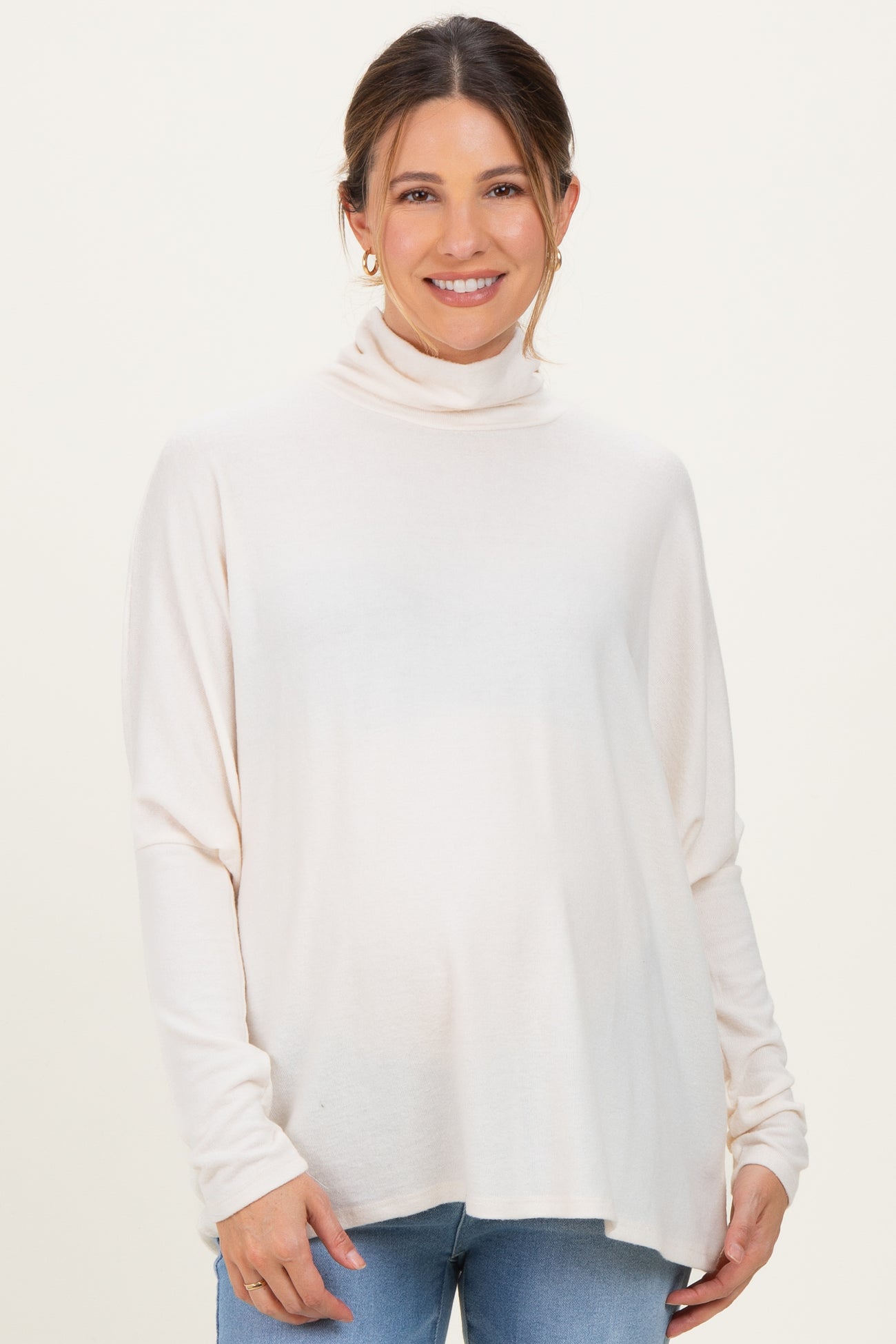 woman wearing a beige maternity turtleneck sweater and blue jeans, showing her baby bump