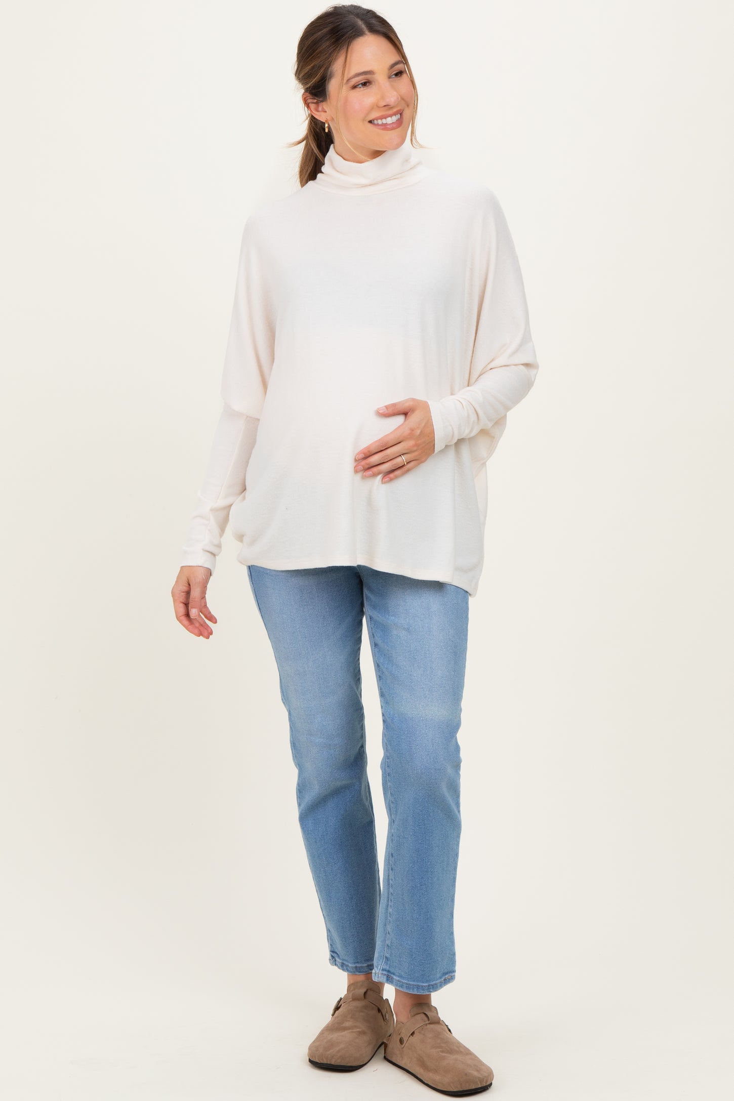 woman wearing a light beige maternity turtleneck top and holding her baby bump, paired with light blue jeans and slippers