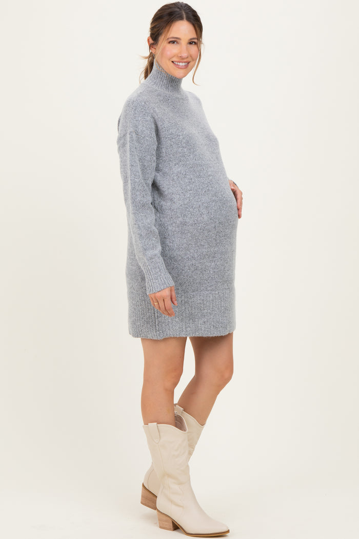 woman wearing a light gray maternity sweater dress with a turtleneck and holding her baby bump, paired with white ankle boots