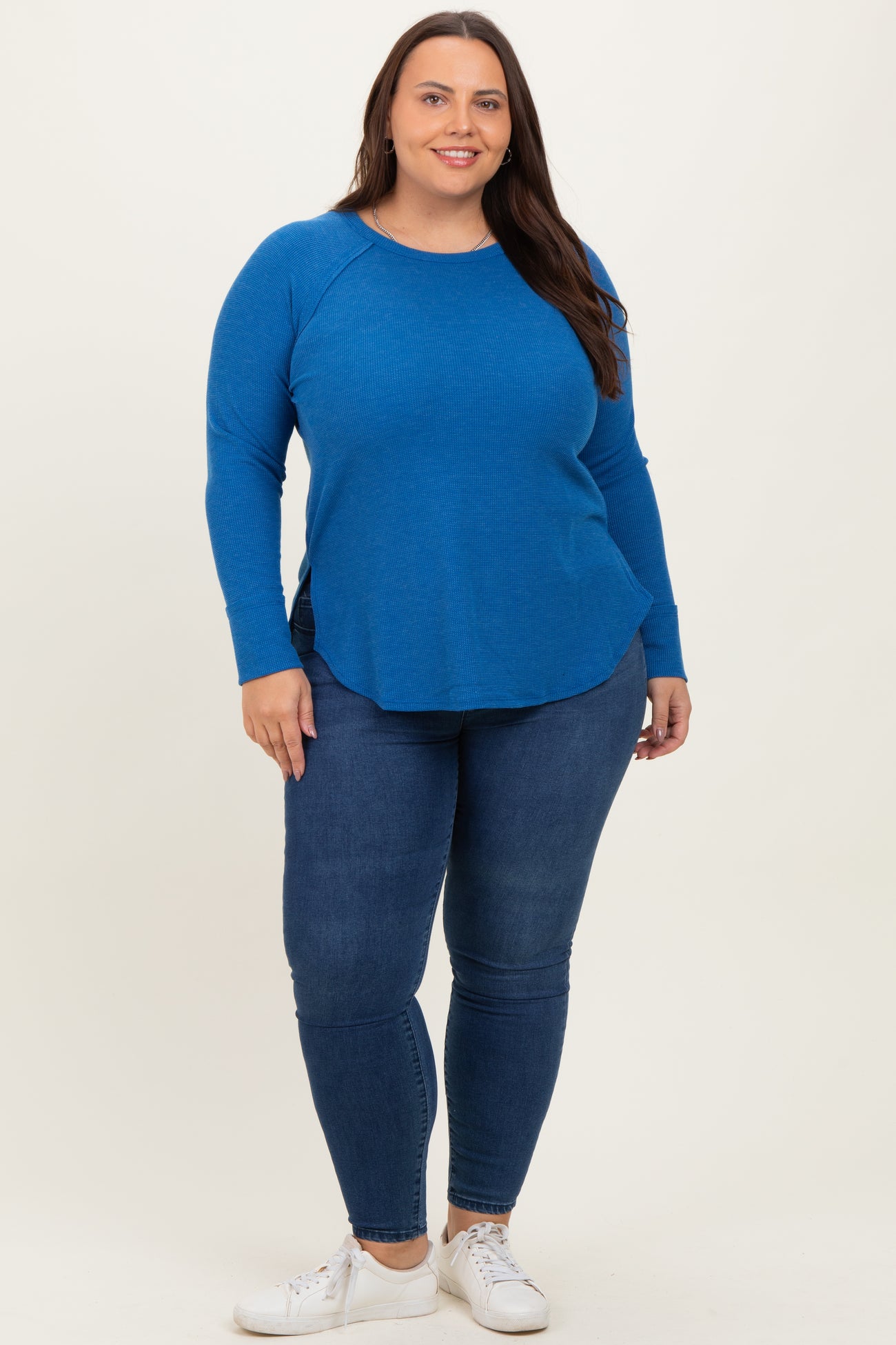 woman wearing a bright blue long sleeve top and dark blue skinny jeans, standing against a neutral background