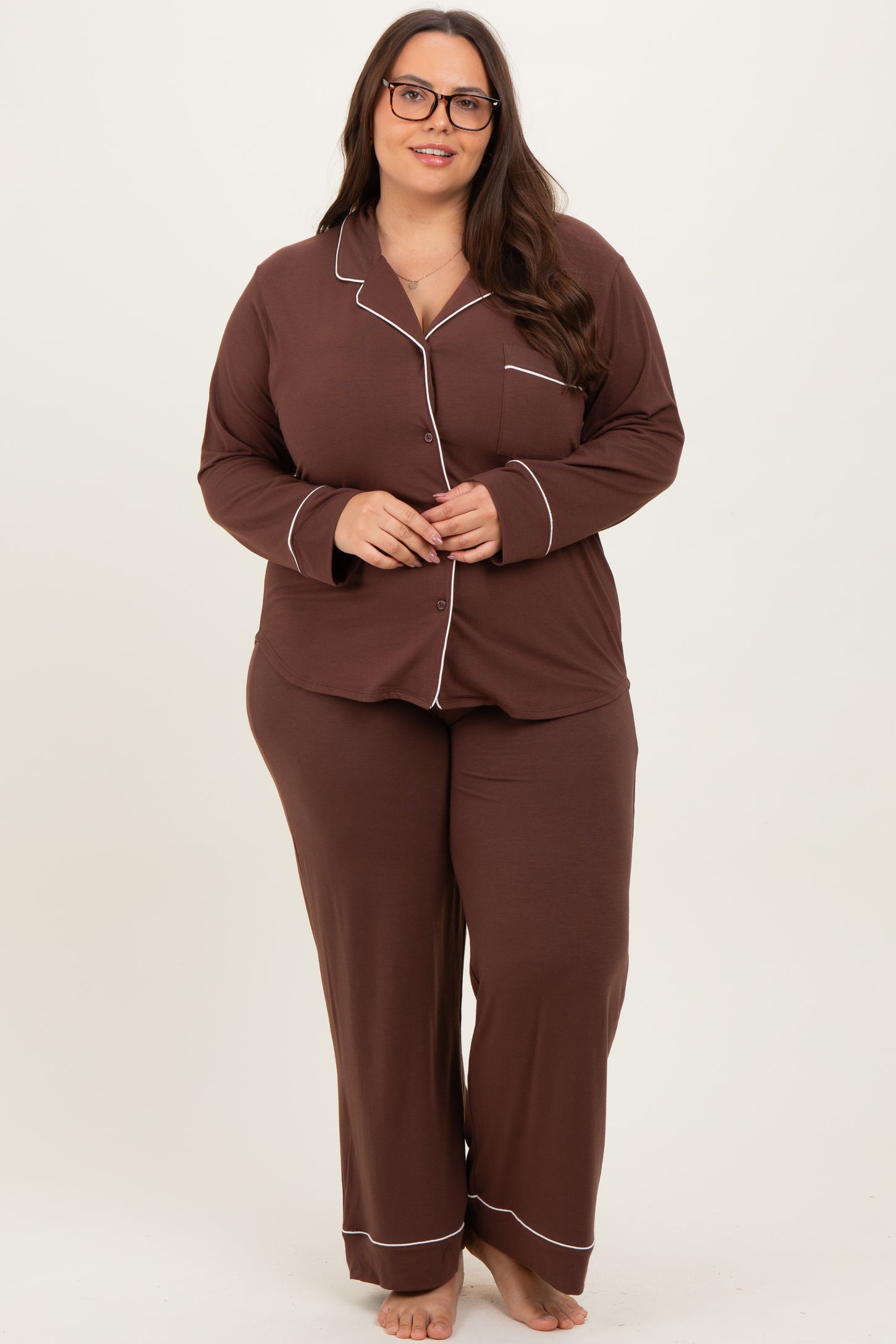 woman wearing a brown plus size pajama set with white piping and long sleeves, standing and smiling