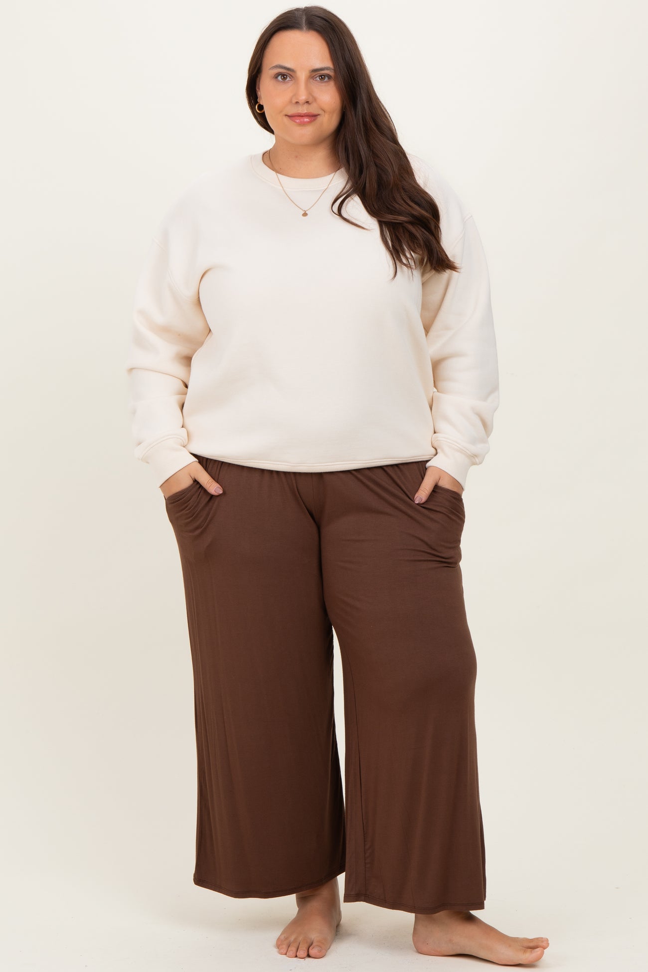 woman wearing a cream oversized sweatshirt and brown wide-leg pants, standing barefoot with hands in pockets