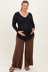 woman wearing a black long sleeve maternity top and brown wide-leg maternity pants, gently holding her baby bump