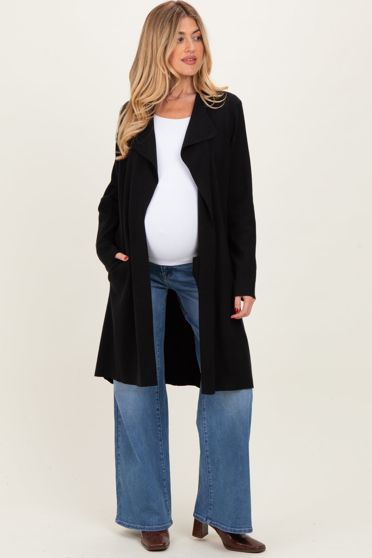 woman wearing a black open-front maternity coat over a white top and blue flared jeans, showing her baby bump