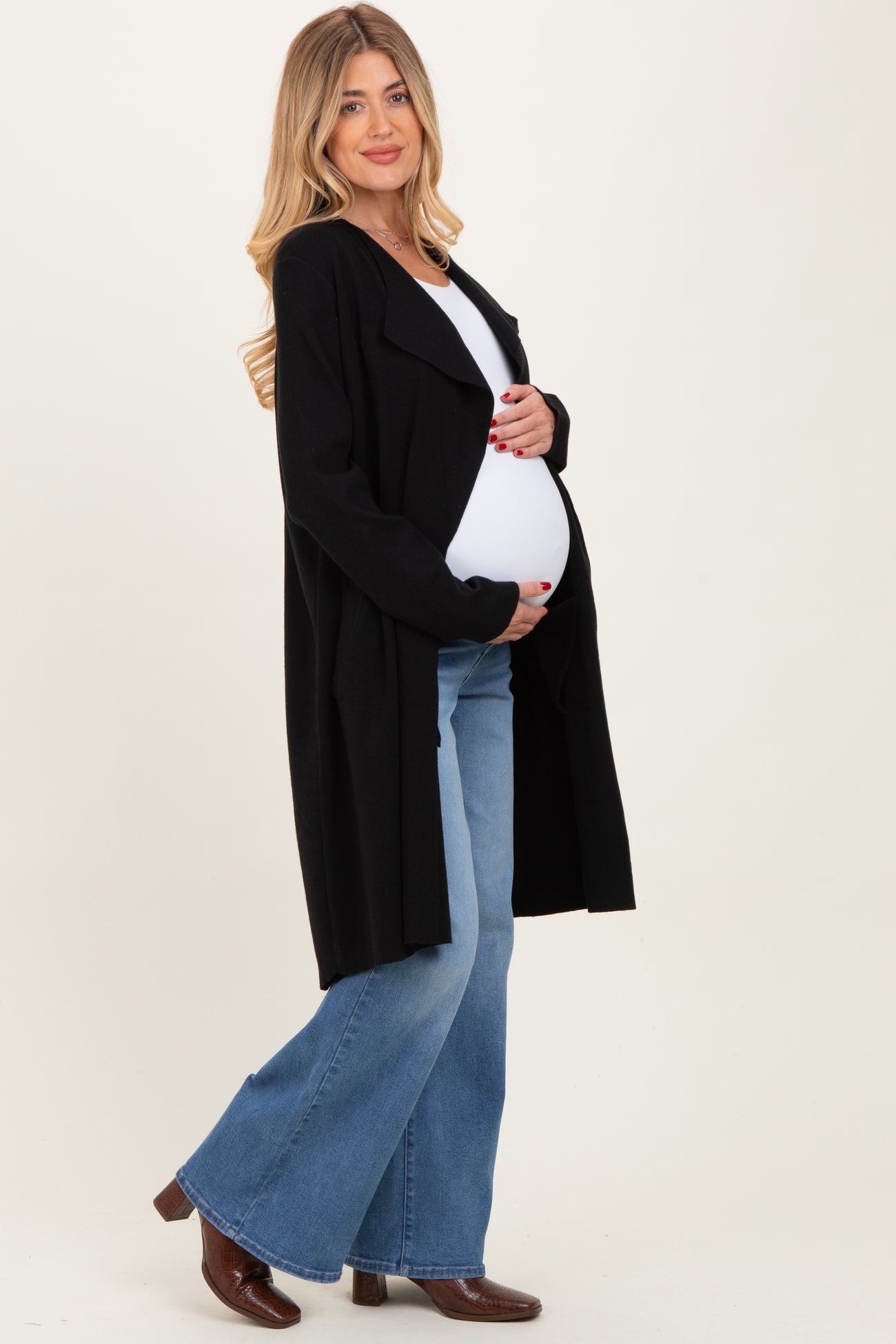 woman wearing a black long maternity cardigan and holding her baby bump while dressed in flared jeans and boots