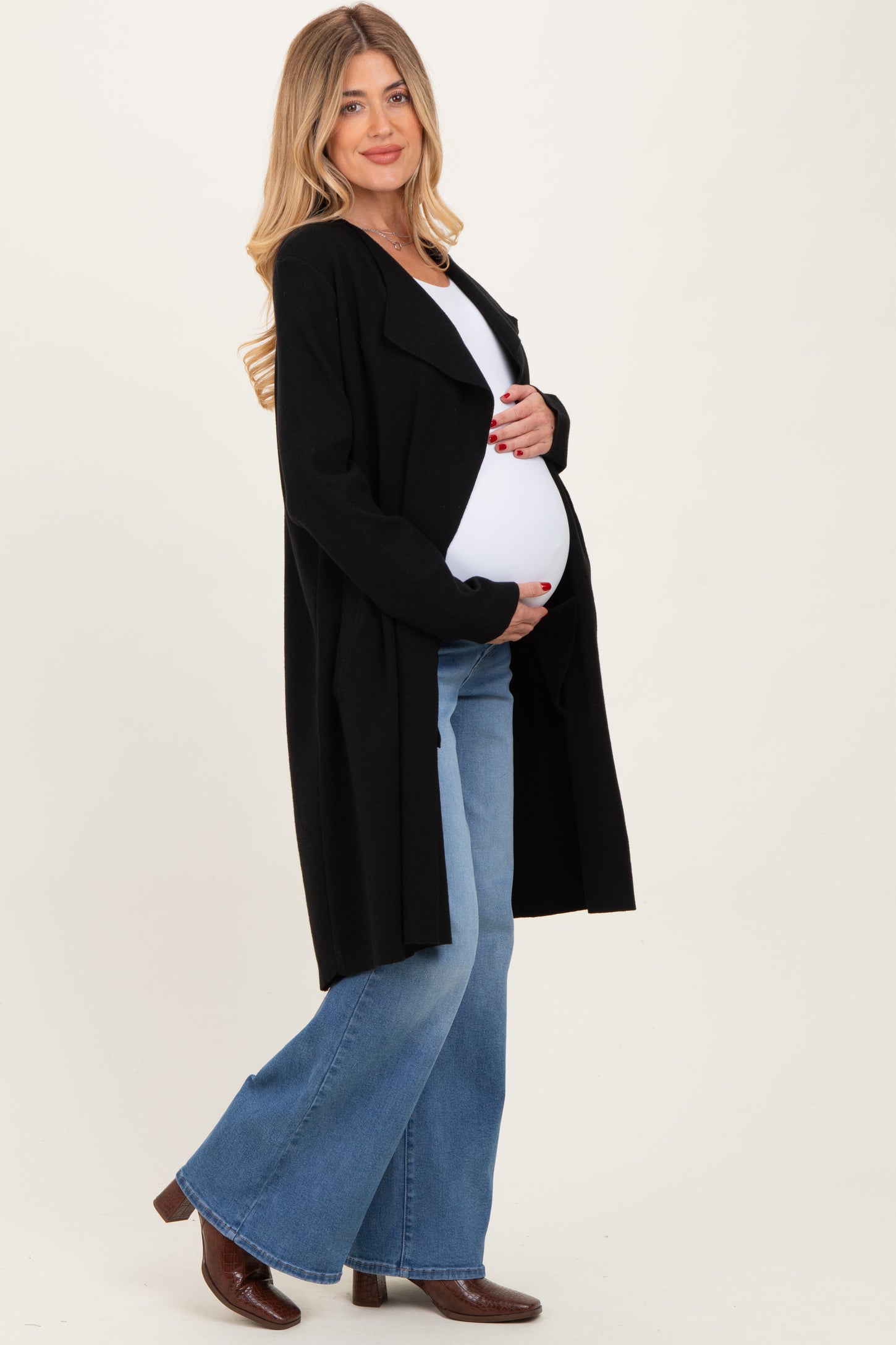 woman wearing a black long maternity cardigan and holding her baby bump while dressed in flared jeans and boots