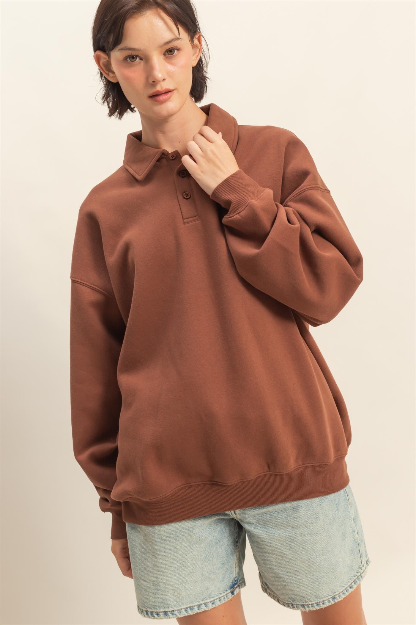 woman wearing a brown oversized polo sweatshirt with a collar and relaxed fit, paired with light blue denim shorts