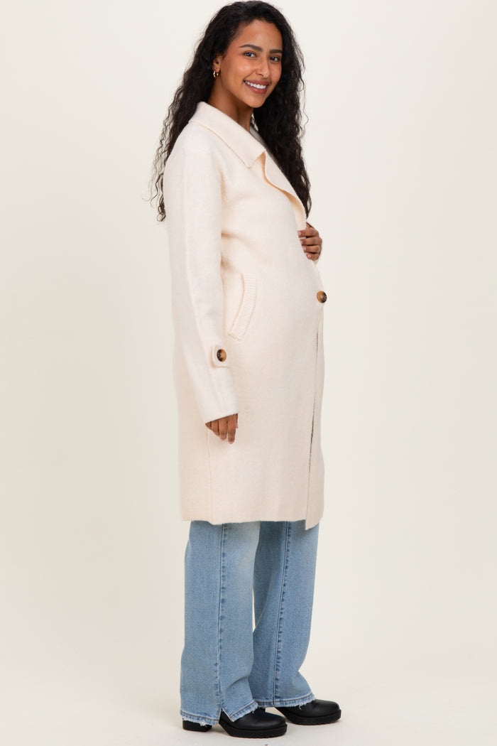 woman wearing a light pink maternity coat with button details and a visible baby bump, paired with blue jeans