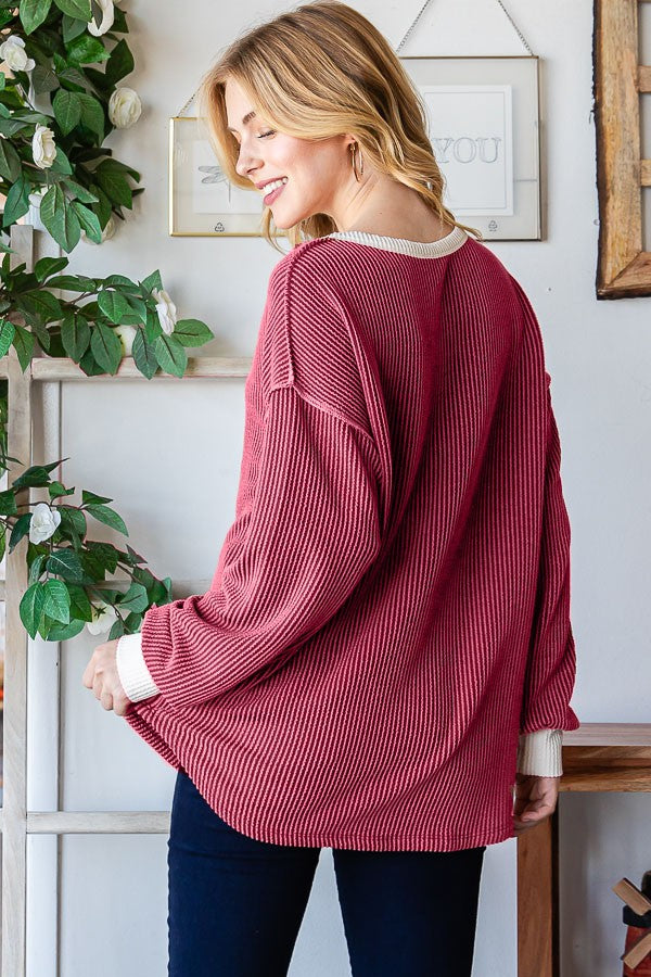 woman wearing a burgundy ribbed oversized top with cream-colored cuffs and back, styled with dark jeans, standing by a plant