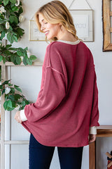 woman wearing a burgundy ribbed oversized top with cream-colored cuffs and back, styled with dark jeans, standing by a plant