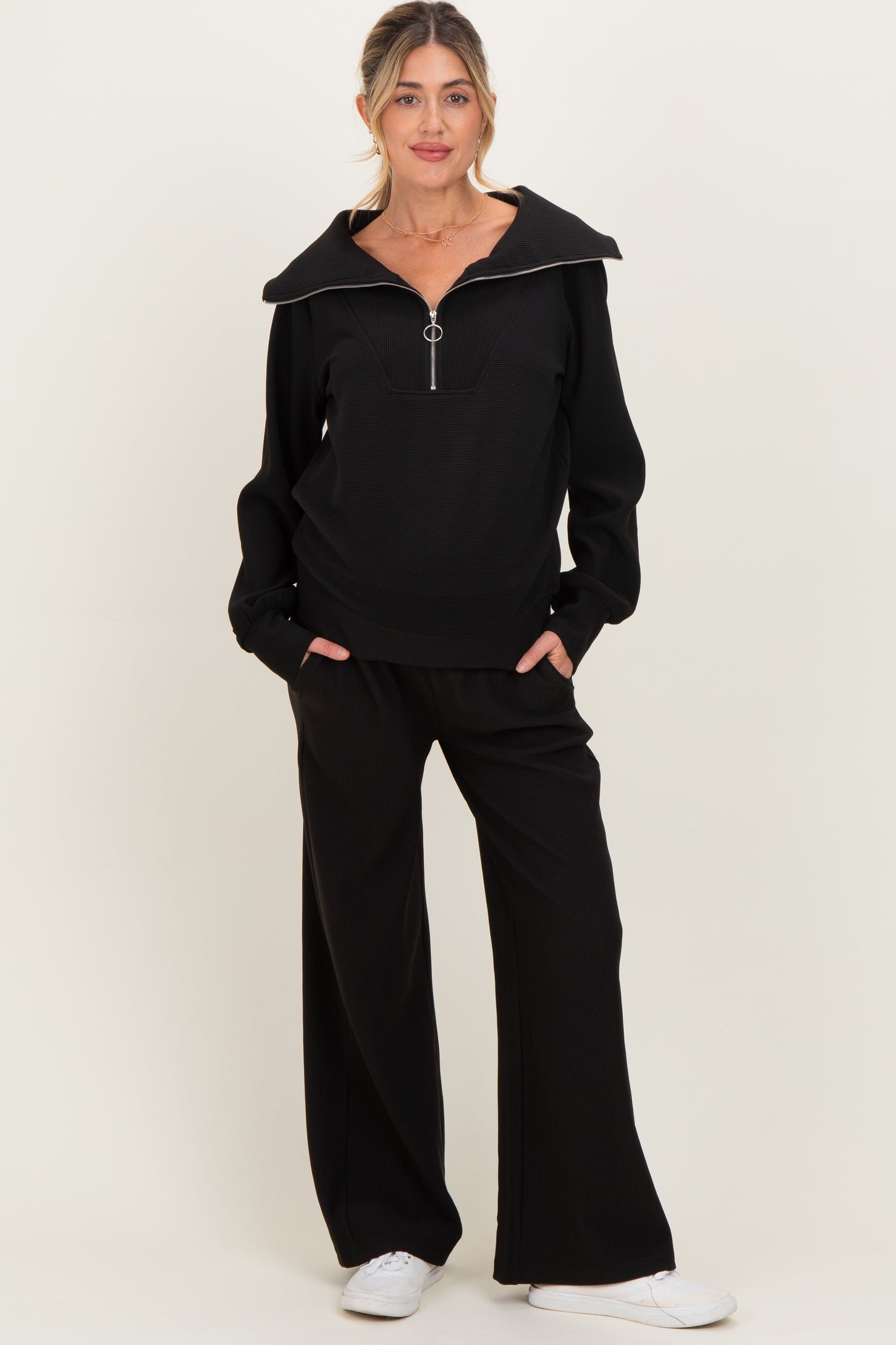 woman wearing a black maternity sweatshirt with a zip-up collar and matching wide-leg pants, styled with white sneakers