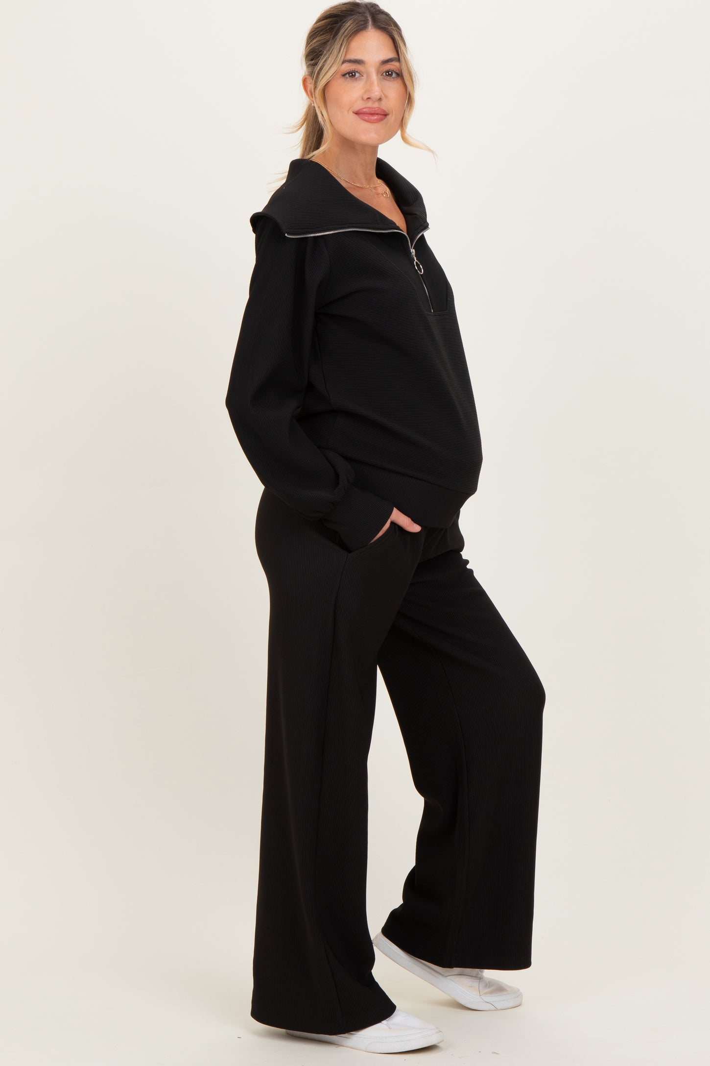 woman wearing a black maternity zip-up sweatshirt with a collar and matching wide-leg pants, holding her baby bump