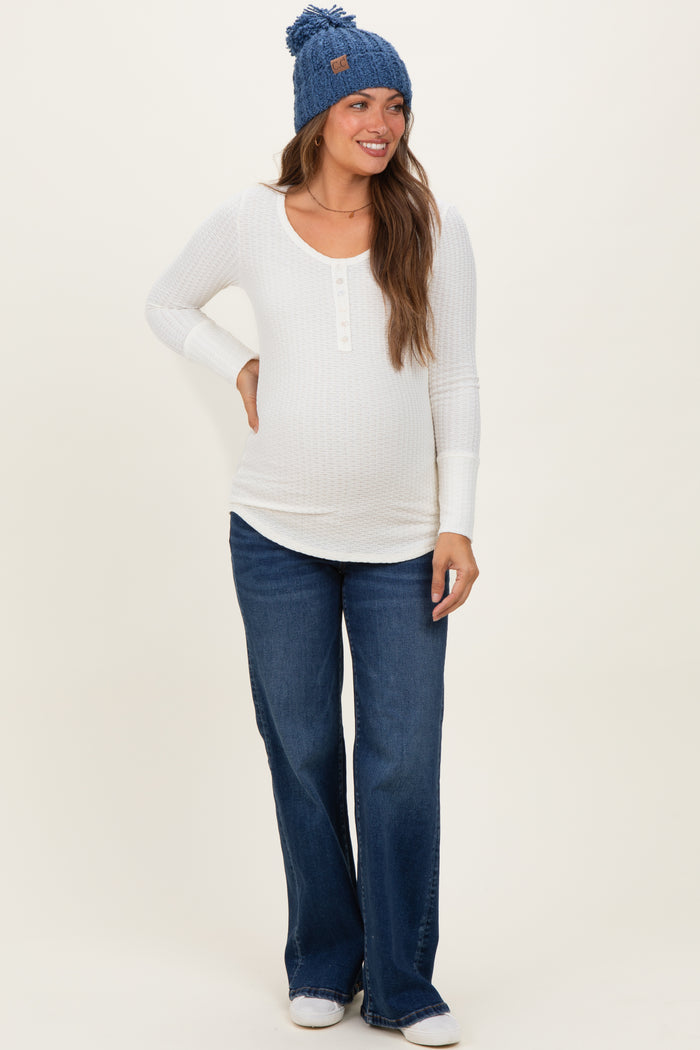 woman wearing a white maternity long sleeve henley top, flared denim jeans, and a blue beanie with a baby bump visible