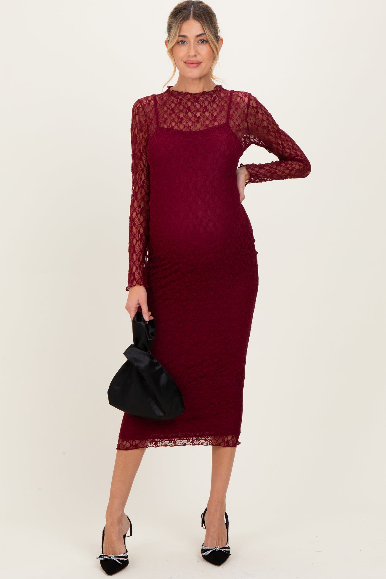 woman wearing a burgundy lace maternity dress with long sleeves and a fitted silhouette, holding a black handbag and showing a baby bump