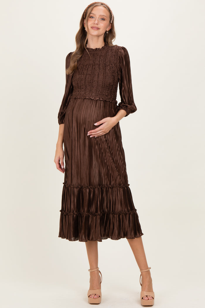 woman wearing a brown pleated maternity dress with smocked bodice and ruffled hem, cradling her baby bump