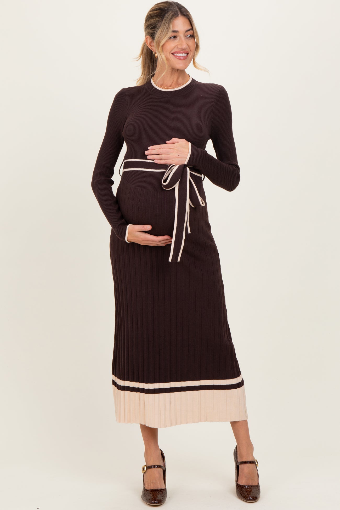 woman wearing a dark brown ribbed maternity dress with a tie waist and light beige hem, holding her baby bump