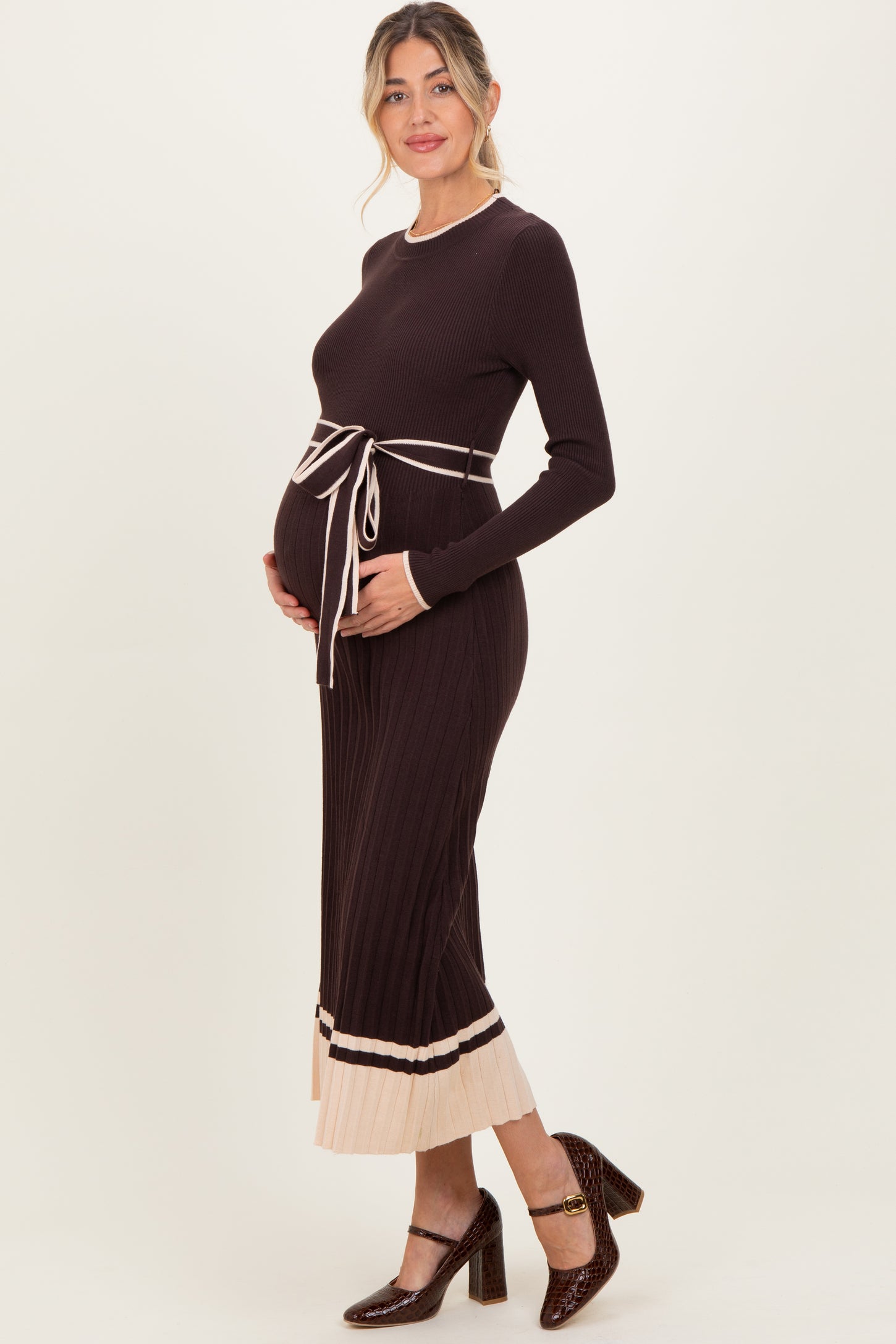 woman wearing a dark brown ribbed maternity dress with a beige tie waist and contrasting hem, gently cradling her baby bump