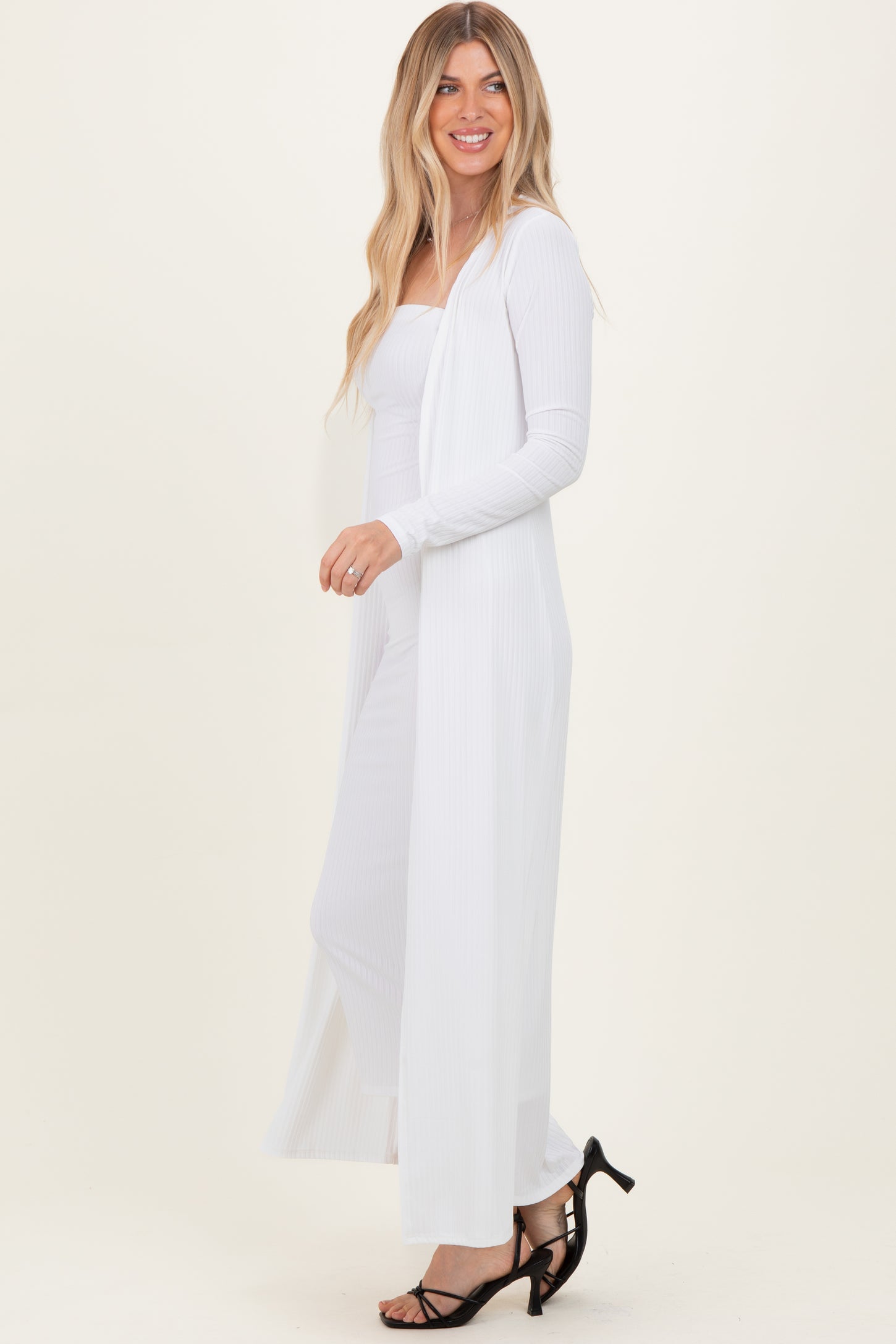 woman wearing a long white ribbed duster cardigan over a matching fitted dress and black strappy heels