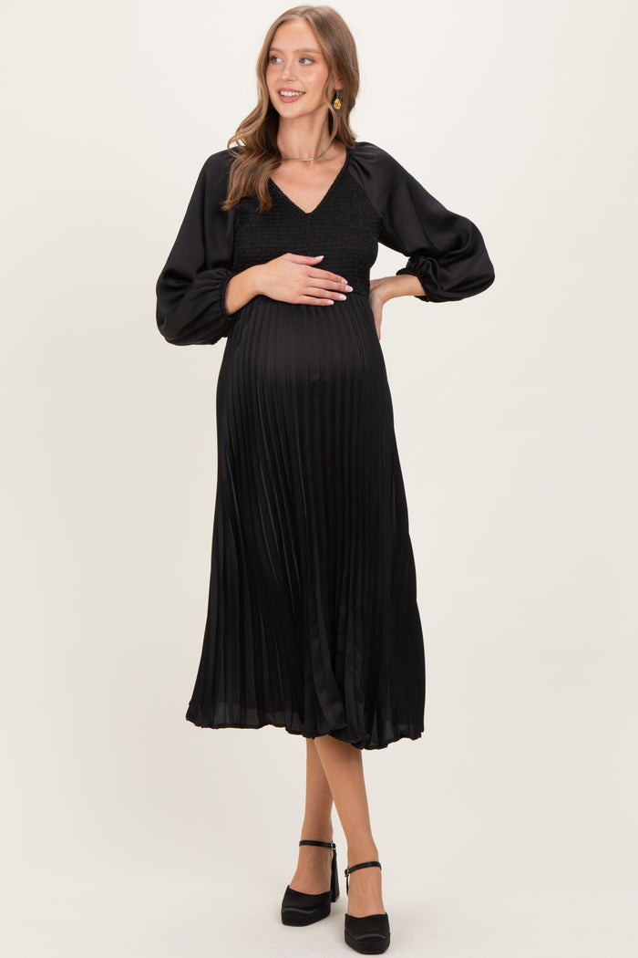 woman wearing a black pleated maternity dress with a v-neck and puff sleeves, holding her baby bump