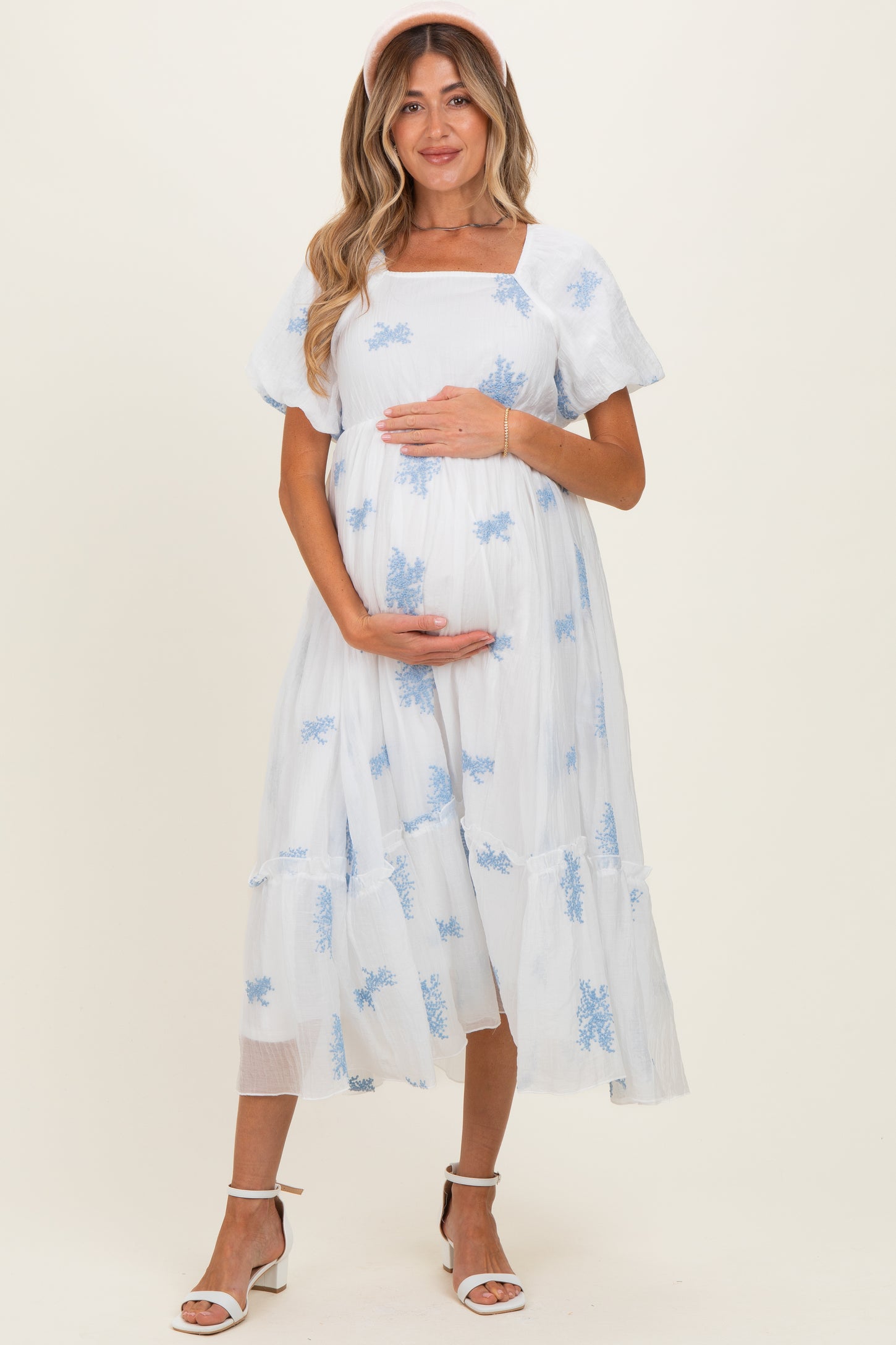 woman wearing a light blue maternity dress with embroidered floral details and short puff sleeves, holding her baby bump