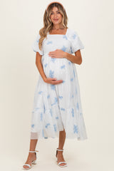 woman wearing a light blue maternity dress with embroidered floral details and short puff sleeves, holding her baby bump