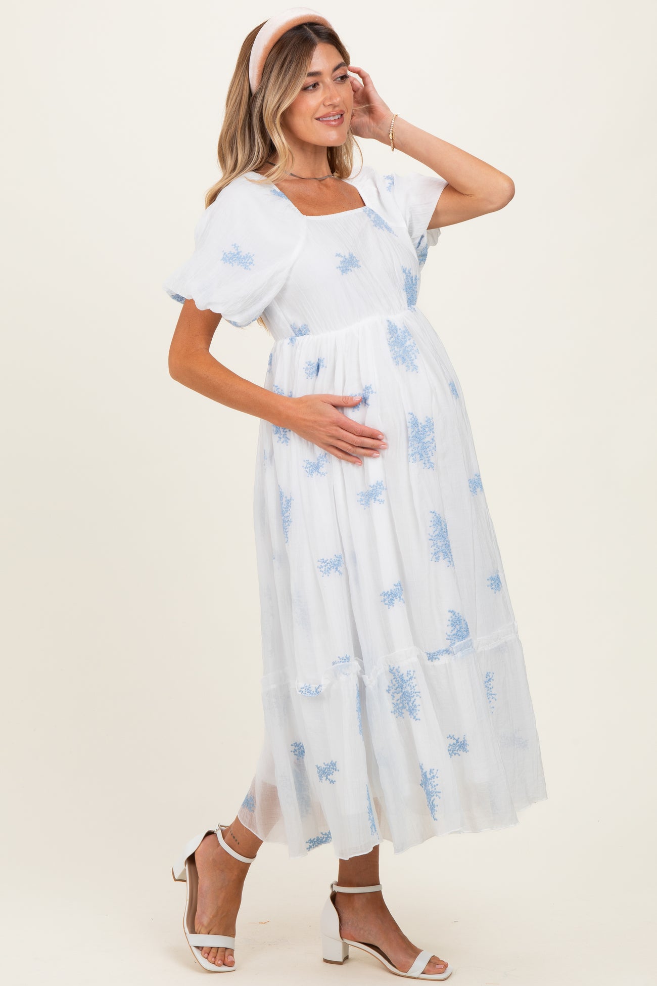 woman wearing a white maternity dress with blue floral prints and short puff sleeves, holding her baby bump