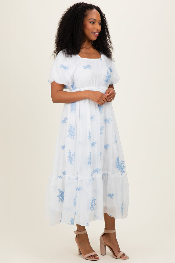 woman wearing a white midi dress with blue floral embroidery and puff sleeves, standing in profile