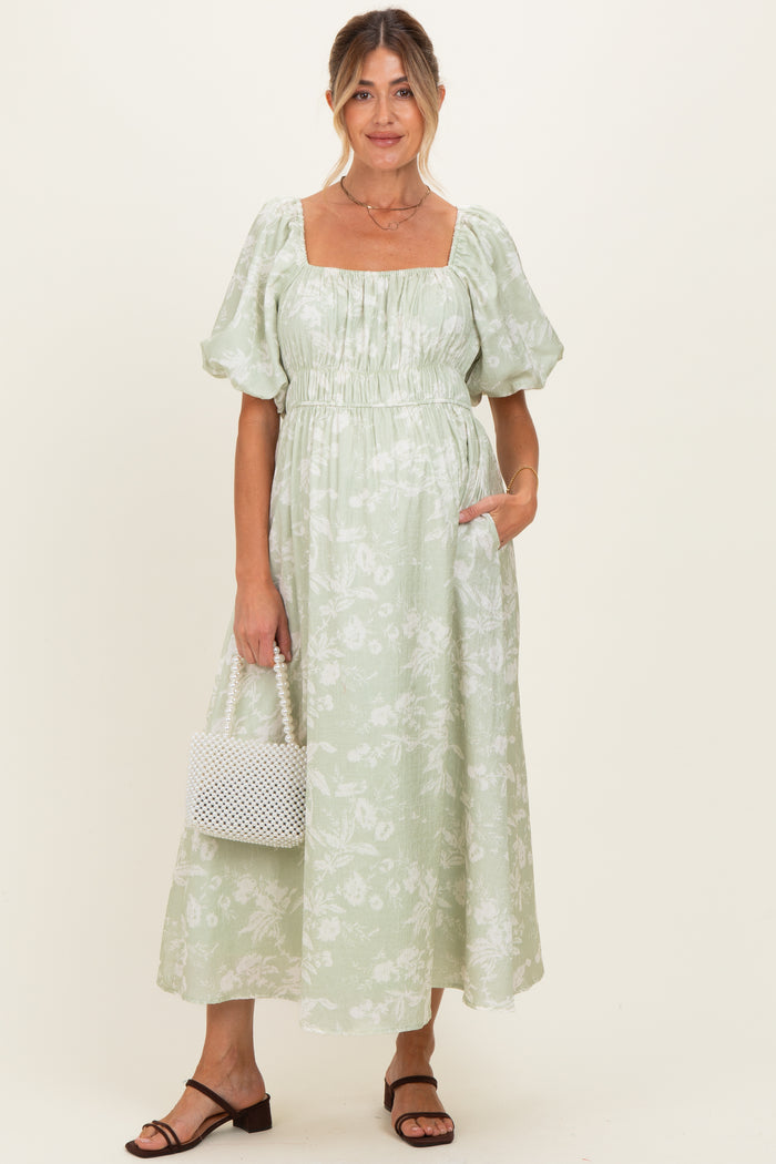 woman wearing a light green floral maternity dress with puffed sleeves and pockets, holding a white bag