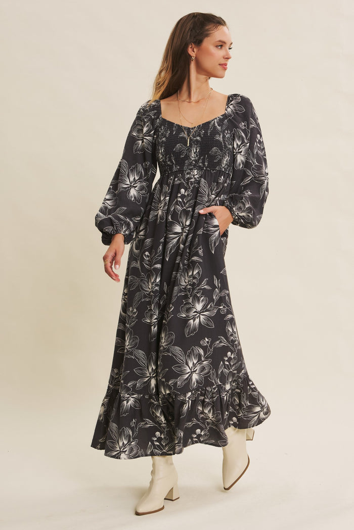 woman wearing a black floral maxi dress with puff sleeves and a tiered hem, posing with hands in pockets
