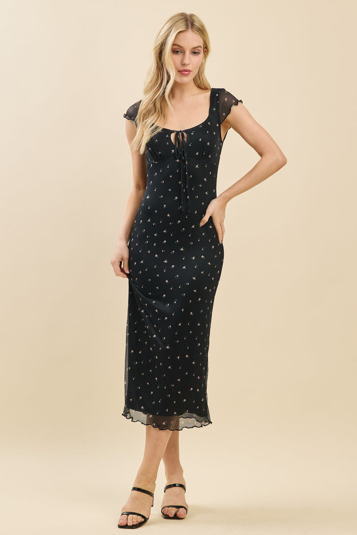woman wearing a black midi dress with floral print and a lace-up neckline, standing in a relaxed pose