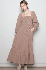 woman wearing a brown checkered long sleeve maxi dress with a smocked bodice and tiered ruffle hem, posing with hands in pockets