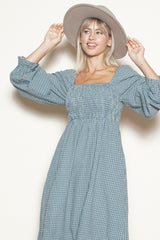 woman wearing a blue checkered long sleeve dress with a square neckline and puffed sleeves, holding a wide-brim hat