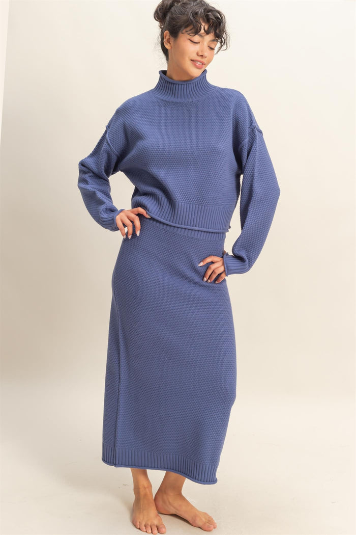 woman wearing a knit blue long sleeve turtleneck sweater and matching maxi skirt, standing barefoot