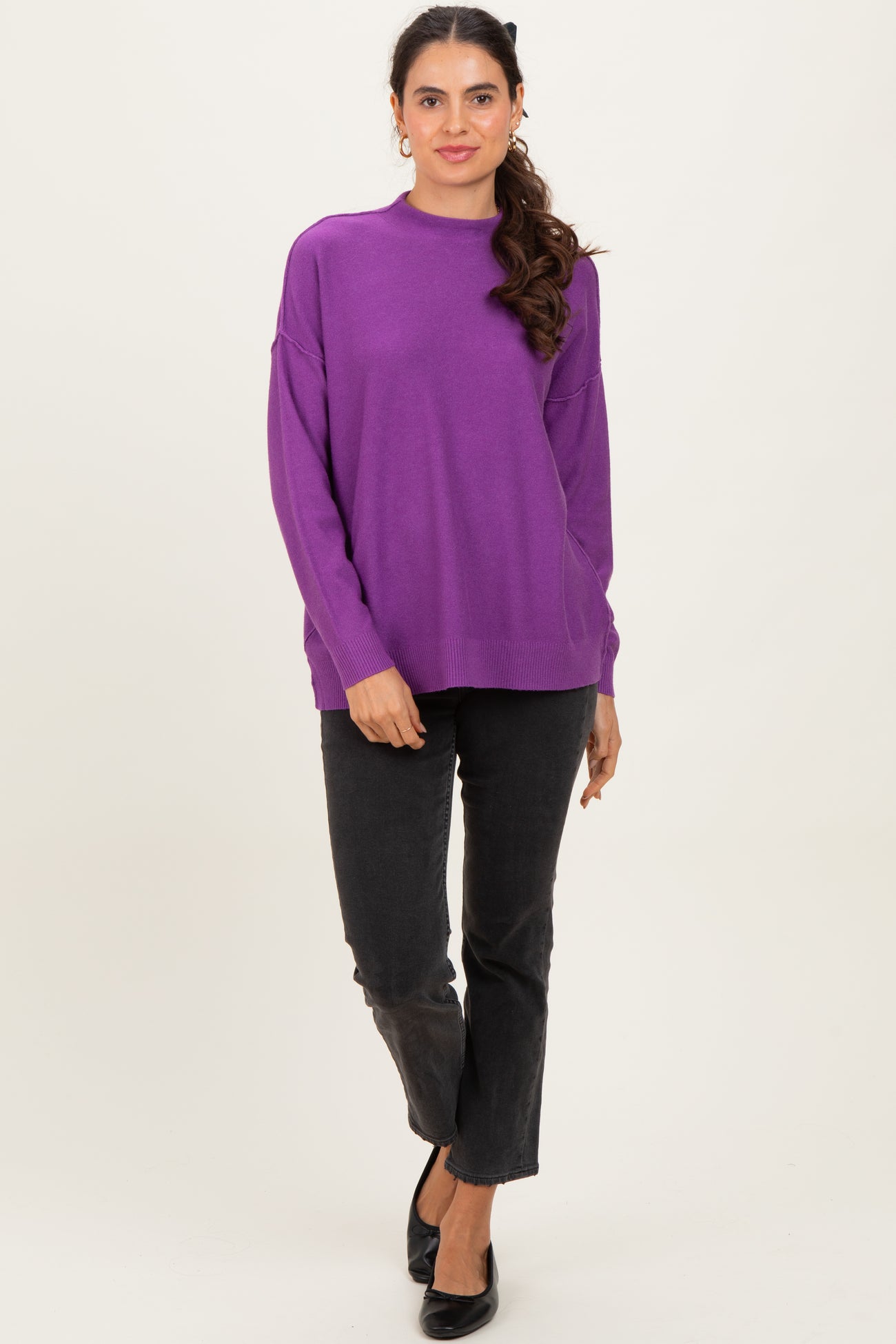 woman wearing a purple oversized sweater with long sleeves and black pants, standing and smiling