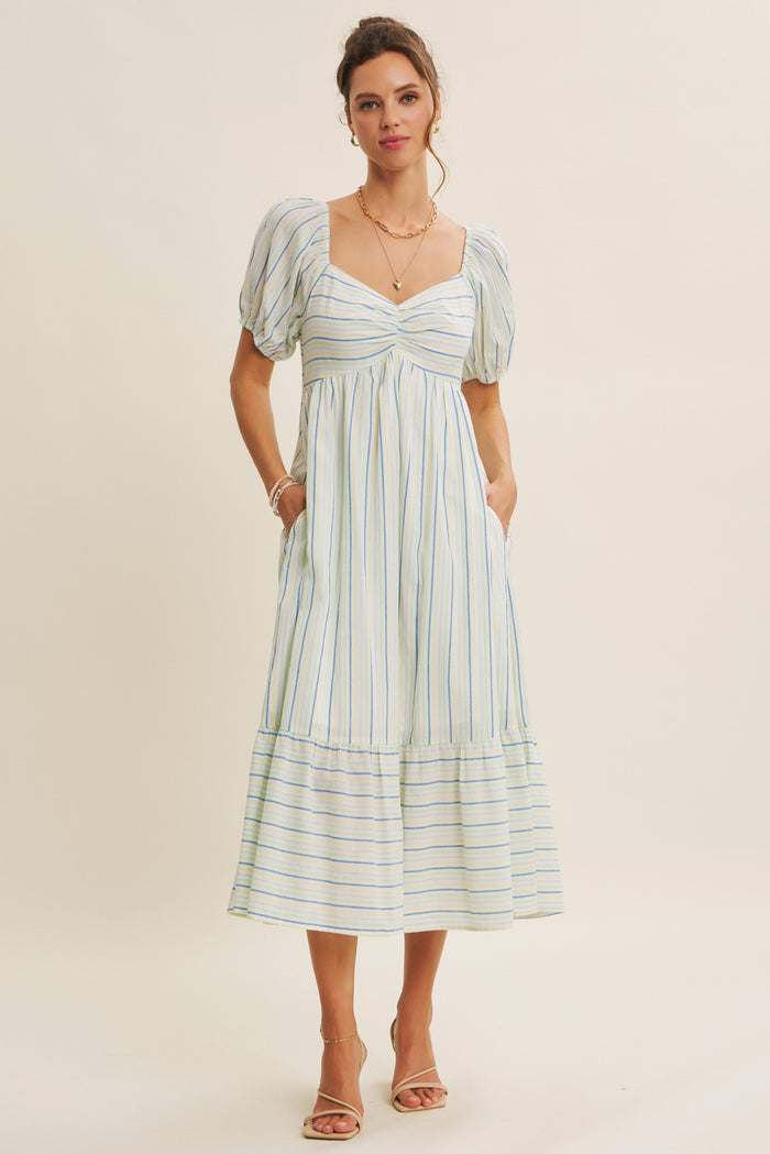 woman wearing a light blue striped midi dress with puff sleeves and a tiered hem, standing with pockets visible