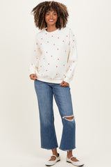 woman wearing a cream maternity sweatshirt with red hearts and blue ripped jeans, smiling and standing with hands on her hips