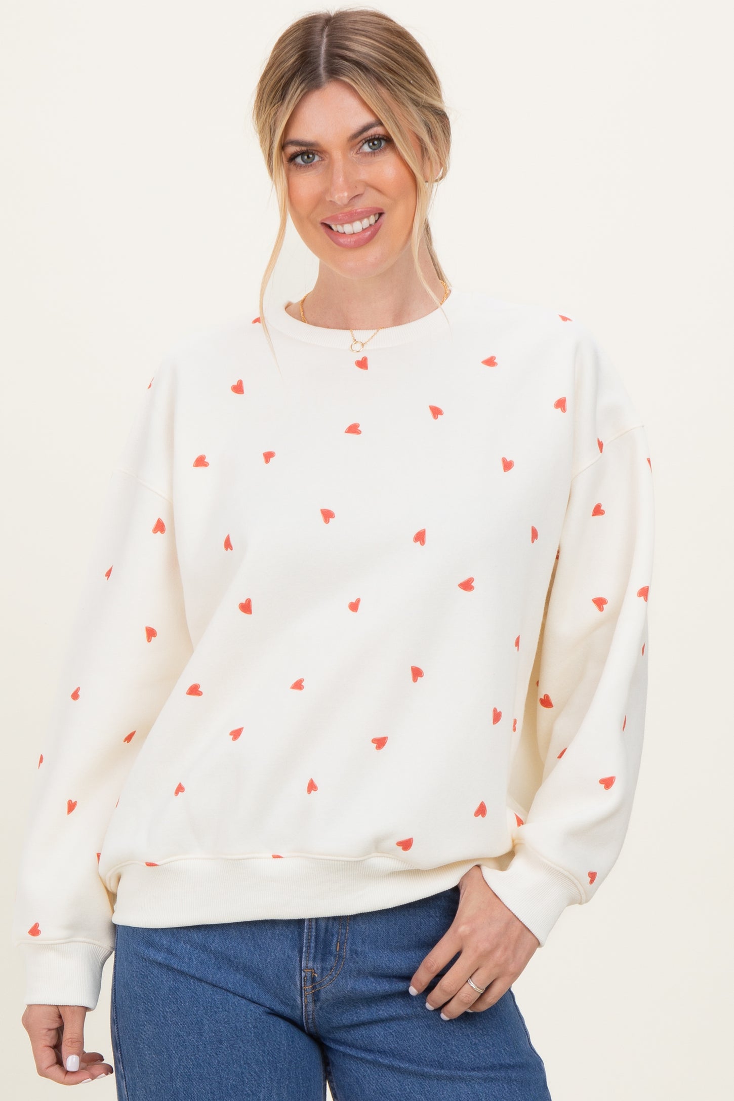 woman wearing a cream sweatshirt with red heart print and relaxed fit, smiling while posing with hands in front