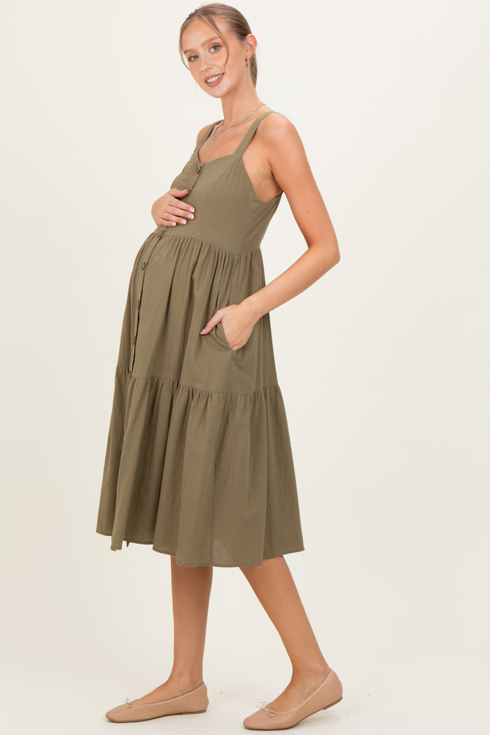 woman wearing an olive green maternity dress with adjustable straps, a tiered skirt, and pockets, showcasing her baby bump