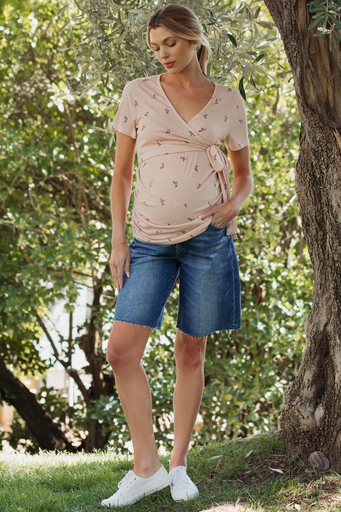 woman wearing a pink floral maternity wrap top and denim bermuda shorts, with a visible baby bump