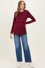 woman wearing a burgundy ribbed maternity top with a scalloped hem and a visible baby bump paired with blue wide leg jeans