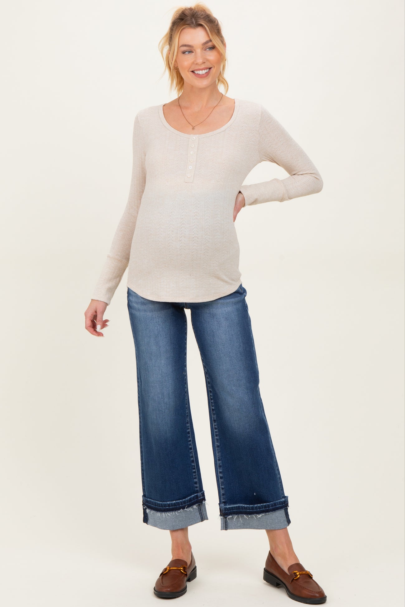 woman wearing a beige maternity long sleeve top and blue wide-leg jeans, showing her baby bump
