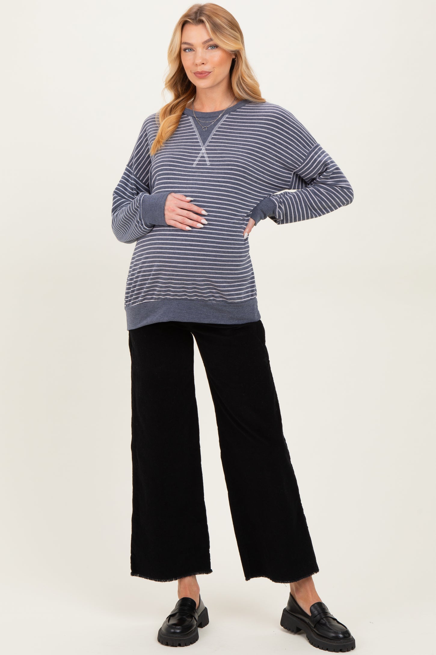 woman wearing a navy and white striped maternity sweatshirt and black wide-leg pants, softly cradling her baby bump