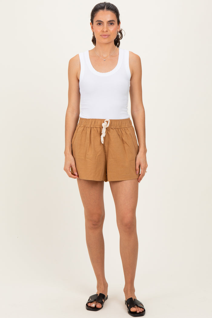 woman wearing a white tank top and brown drawstring shorts, standing against a neutral background