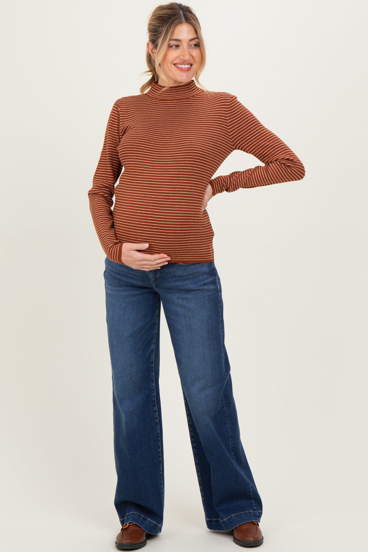 woman wearing a rust striped maternity long sleeve top and blue flared jeans, holding her baby bump
